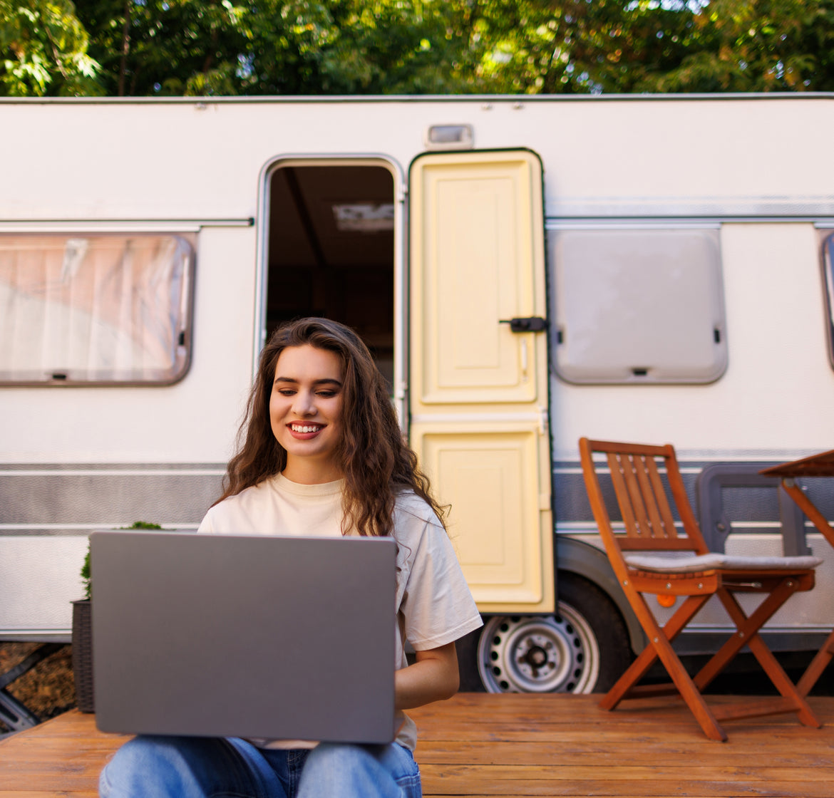 A man works on a laptop under an RV awning at sunset beside the water